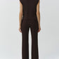 A person stands facing away, wearing the Simkhai Maple Top—a sleeveless knit brown turtleneck sweater—with brown high-waisted pants against a plain light background.