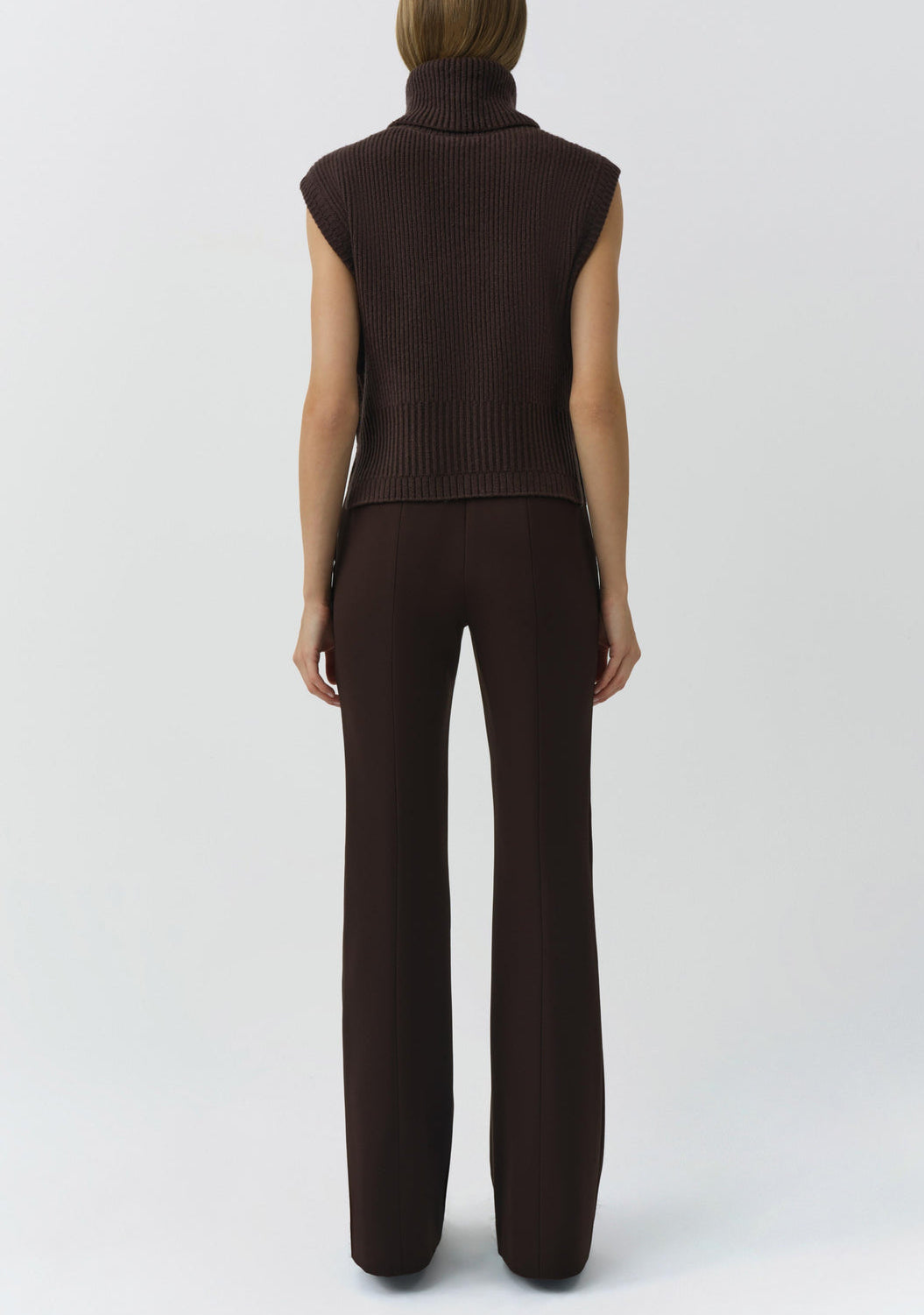 A person stands facing away, wearing the Simkhai Maple Top—a sleeveless knit brown turtleneck sweater—with brown high-waisted pants against a plain light background.