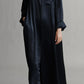 A person wearing TWP Jenny's Silk Gown, a long, dark blue maxi shirt dress with long sleeves and black shoes. The relaxed silhouette features a pocket on the left chest and a collar.