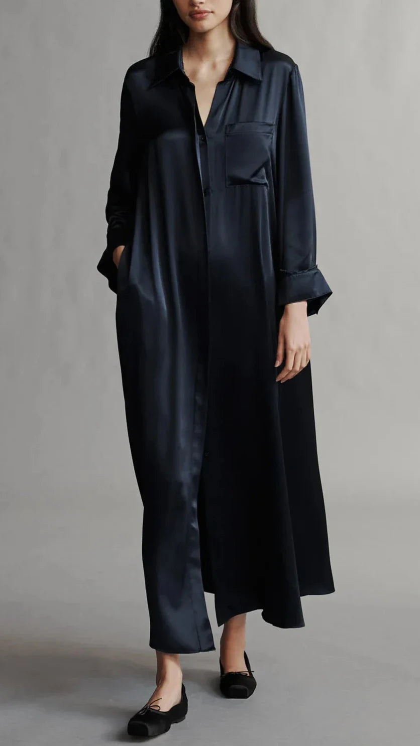 A person wearing TWP Jenny's Silk Gown, a long, dark blue maxi shirt dress with long sleeves and black shoes. The relaxed silhouette features a pocket on the left chest and a collar.