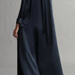 A person wearing TWP's Jenny's Silk Gown in a long, dark hue with a relaxed silhouette is standing with their back turned. They have one arm bent at the elbow and are wearing black flats.