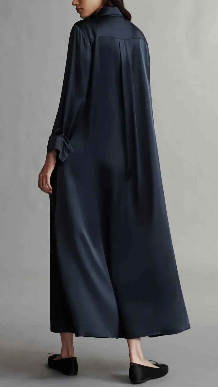 A person wearing TWP's Jenny's Silk Gown in a long, dark hue with a relaxed silhouette is standing with their back turned. They have one arm bent at the elbow and are wearing black flats.