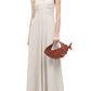 A woman stands against a white background wearing the Staud Madison Linen Dress—sleeveless, floor-length with a sweetheart neckline—and holds a brown shark-shaped handbag.