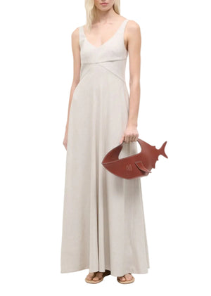 A woman stands against a white background wearing the Staud Madison Linen Dress—sleeveless, floor-length with a sweetheart neckline—and holds a brown shark-shaped handbag.