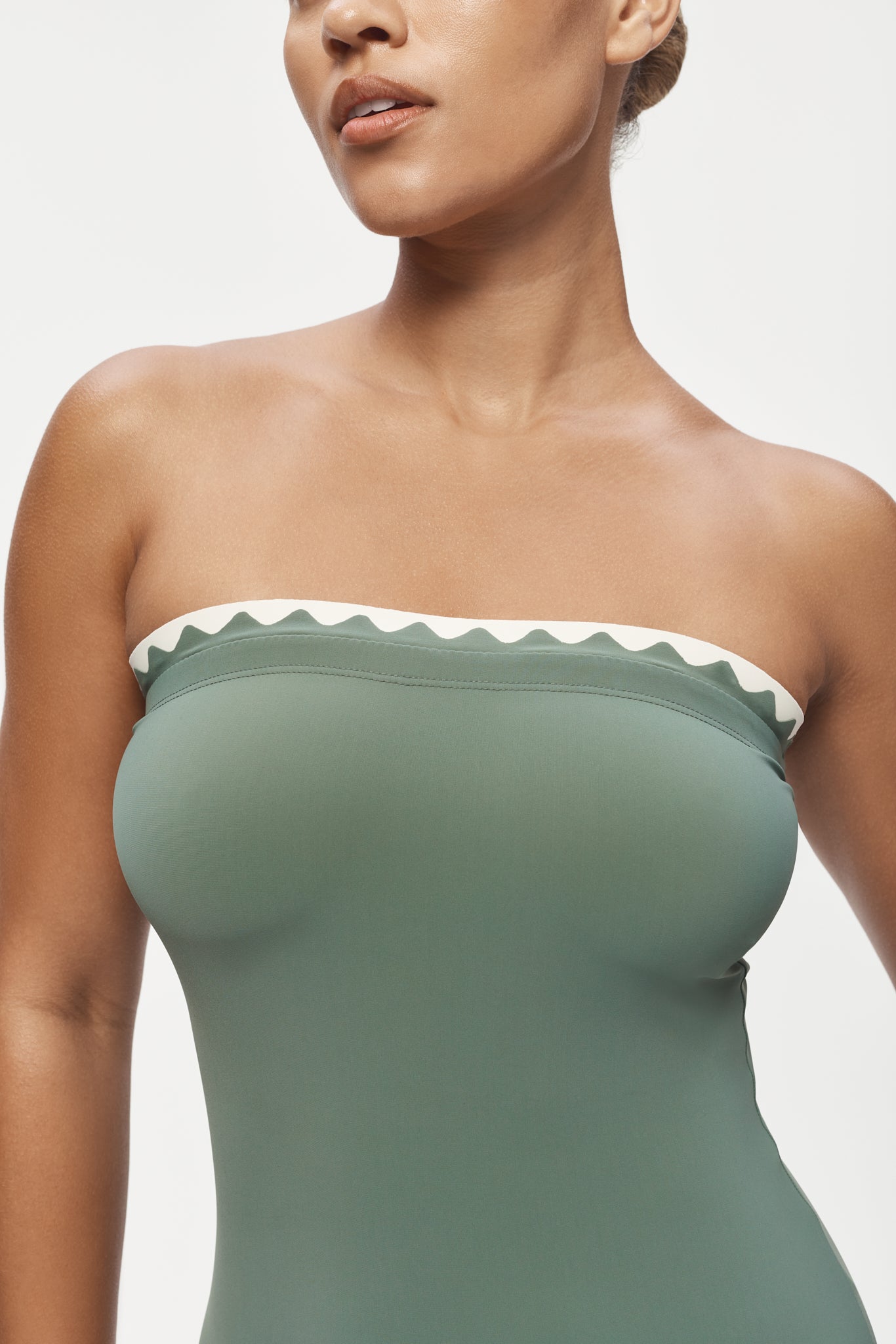A woman models the Karla Colletto Lia Bandeau One Piece in sage green with a white rickrack trim, shown from shoulders to mid-torso against a plain background.