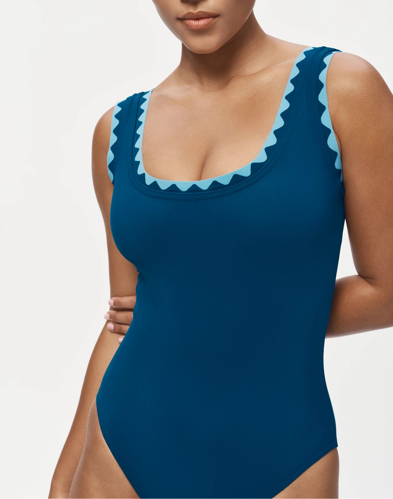 A woman wears the Karla Colletto Lia Round Neck One Piece, featuring blue fabric with rickrack trim and light blue zigzag details along the neckline and shoulder straps.