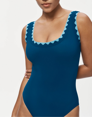 A woman wears the Karla Colletto Lia Round Neck One Piece, featuring blue fabric with rickrack trim and light blue zigzag details along the neckline and shoulder straps.