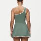 A woman stands facing away in the Karla Colletto Lia Skirt, a comfort-first green one-shoulder swimsuit dress featuring white rickrack trim and an A-line skirt, set against a plain light background.