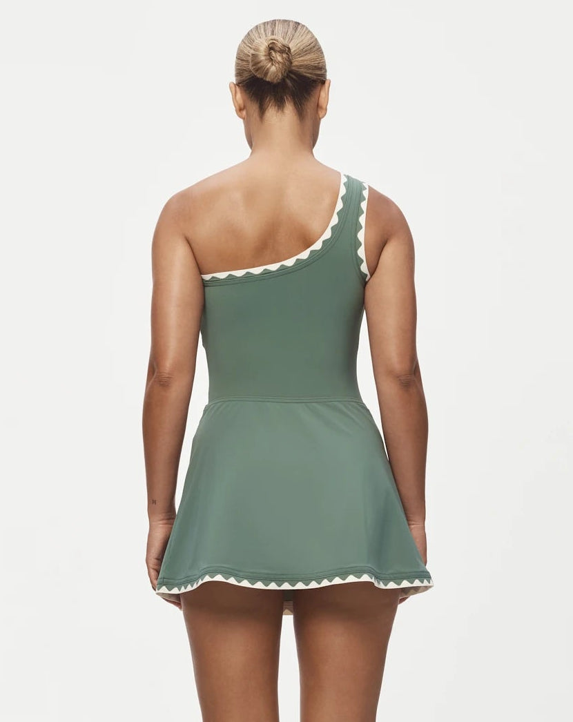 A woman stands facing away in the Karla Colletto Lia Skirt, a comfort-first green one-shoulder swimsuit dress featuring white rickrack trim and an A-line skirt, set against a plain light background.