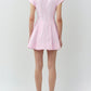 A woman faces away, wearing the Simkhai Clemmie Mini Dress in light pink cotton with cap sleeves and a flared skirt, paired with white strappy heels against a plain background.