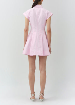 A woman faces away, wearing the Simkhai Clemmie Mini Dress in light pink cotton with cap sleeves and a flared skirt, paired with white strappy heels against a plain background.