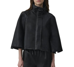 A person wearing the Simkhai Orvik Leather Cape Jacket—a black, high-collar, zip-up design with wide cape sleeves—paired with matching black pants, shown from the waist up.