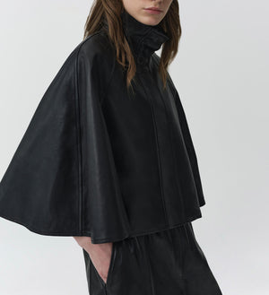 A person wearing the Simkhai Orvik Leather Cape Jacket, featuring dramatic cape sleeves and matching pants, stands against a plain light background.