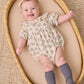A smiling baby wears the Quincy Mae Baby Girls’ Collared Romper, featuring puff sleeves and delicate ruffle accents, paired with a headband and gray knee-high socks, while lying in a woven basket on a textured mat.