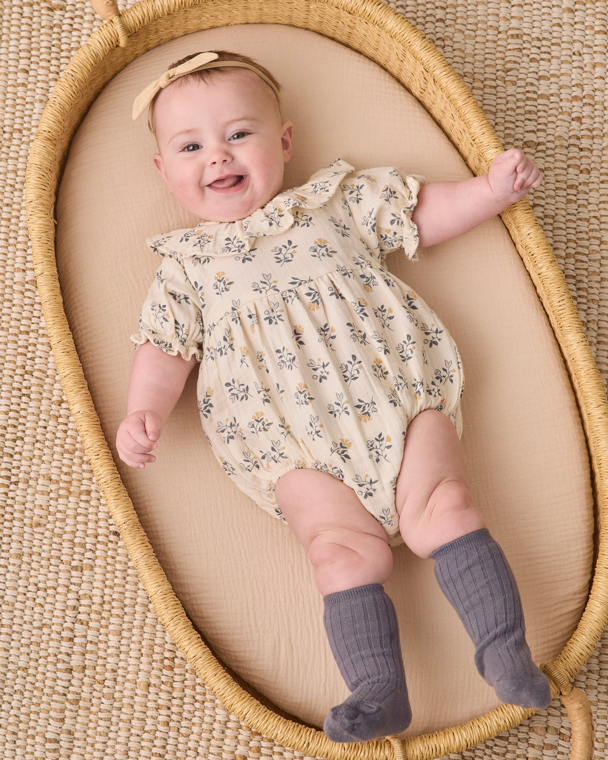 A smiling baby wears the Quincy Mae Baby Girls’ Collared Romper, featuring puff sleeves and delicate ruffle accents, paired with a headband and gray knee-high socks, while lying in a woven basket on a textured mat.