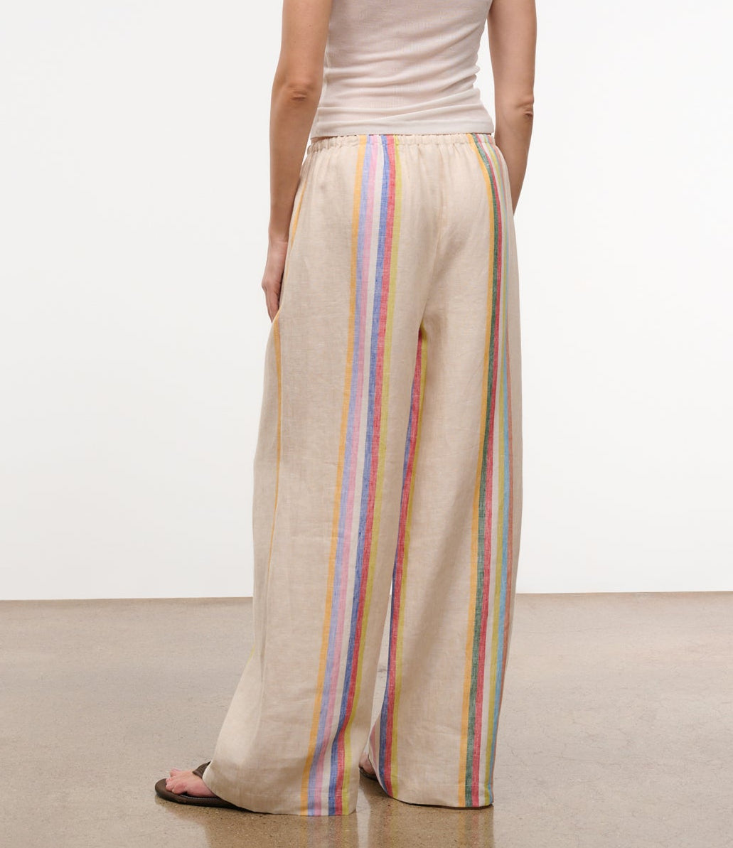 A person wearing Staud's Mateo Linen Pant in relaxed fit with beige vertical multicolored stripes and a light sleeveless top stands on a smooth floor, seen from behind—a stylish warm-weather pick.