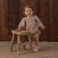A young child wearing the Rylee & Cru Baby Girls’ Adelaide Romper, featuring smocked detailing and puffed sleeves, stands beside a rustic wooden stool on a patterned rug in front of a wooden wall.