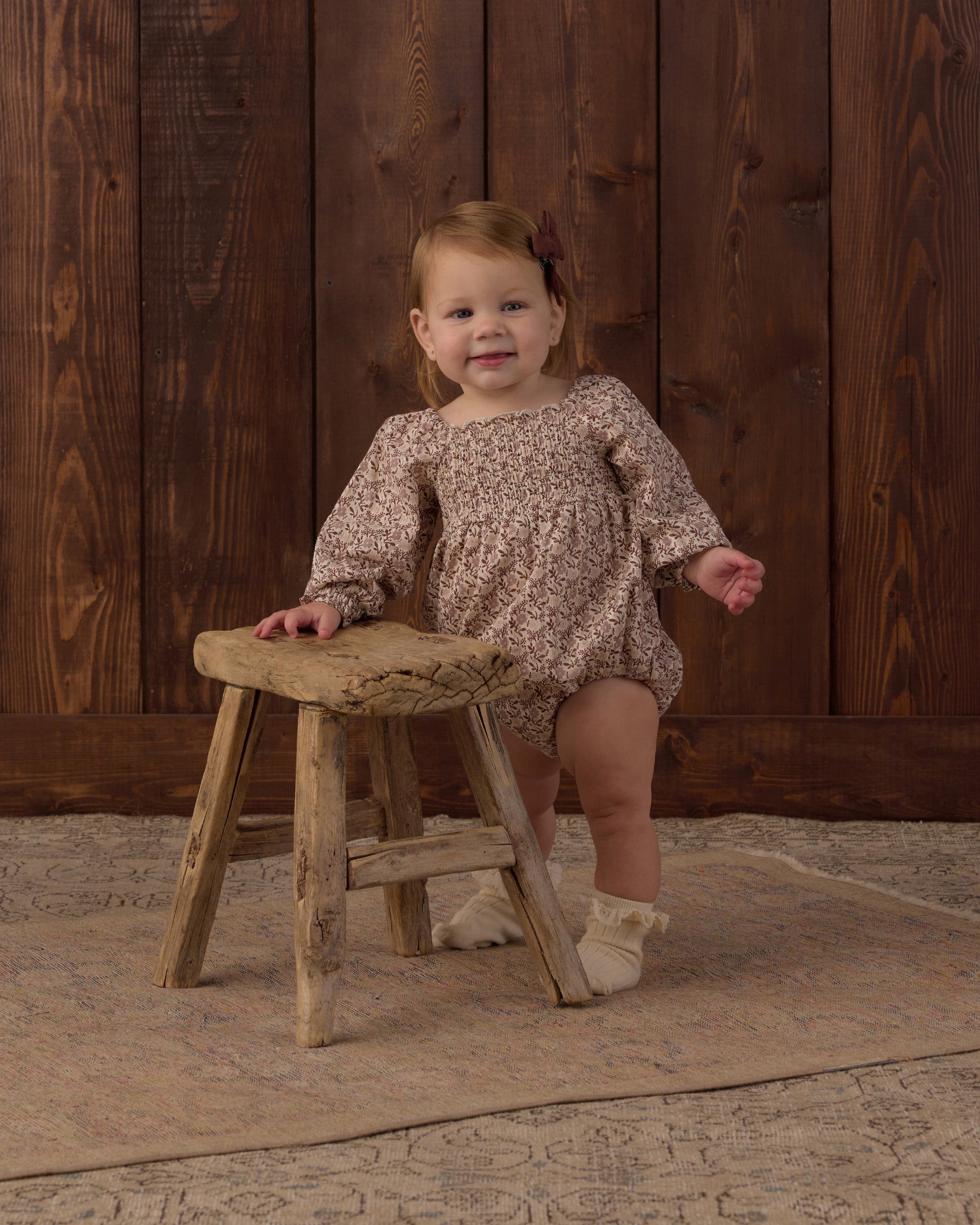 A young child wearing the Rylee & Cru Baby Girls’ Adelaide Romper, featuring smocked detailing and puffed sleeves, stands beside a rustic wooden stool on a patterned rug in front of a wooden wall.