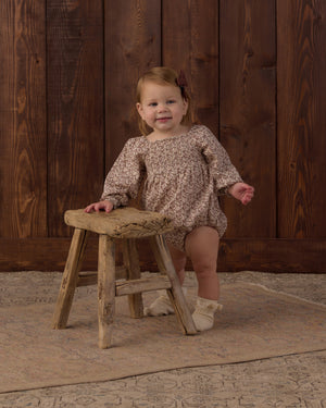 A young child wearing the Rylee & Cru Baby Girls’ Adelaide Romper, featuring smocked detailing and puffed sleeves, stands beside a rustic wooden stool on a patterned rug in front of a wooden wall.