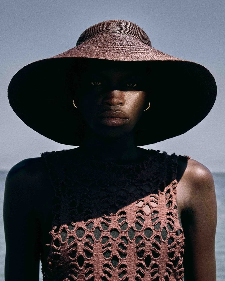 A person stands outdoors by the sea and sky, wearing the Janessa Leone Birdie Visor and a textured sleeveless top.