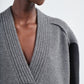 A close-up of a person wearing the Maria McManus Cocoon Cape, an alpaca-wool blend grey knit with a deep V-neck and ribbed detailing, paired with geometric metallic earrings.