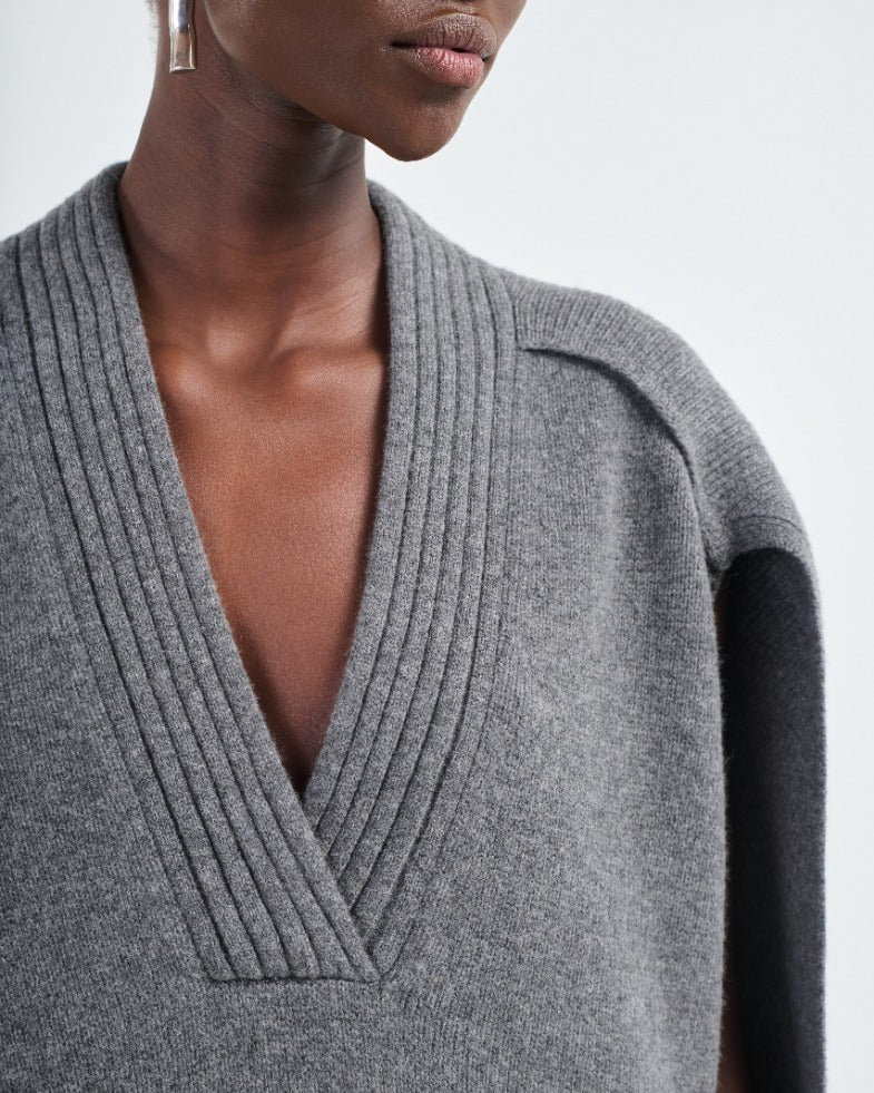 A close-up of a person wearing the Maria McManus Cocoon Cape, an alpaca-wool blend grey knit with a deep V-neck and ribbed detailing, paired with geometric metallic earrings.