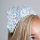 A close-up of a person with blonde hair wearing the Tutu du Monde Snow Queen Tiara, a white beaded headband adorned with large, iridescent floral designs that evoke the look of a delicate flower tiara.
