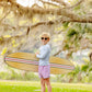 A smiling child in The Beaufort Bonnet Company’s Baby Boys' and Boys' Walker's Wave Swim Shirt with sunglasses stands on grass holding a surfboard, surrounded by trees and greenery.