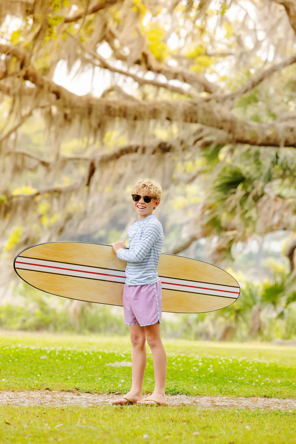 A smiling child in The Beaufort Bonnet Company’s Baby Boys' and Boys' Walker's Wave Swim Shirt with sunglasses stands on grass holding a surfboard, surrounded by trees and greenery.