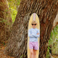 A boy in sunglasses and a striped shirt stands confidently by a tree with his surfboard, sporting The Beaufort Bonnet Company’s Baby Boys' and Boys' Walker's Wave Swim Shirt for stylish beach days and built-in UPF protection.