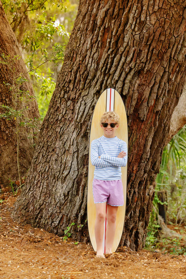 A boy in sunglasses and a striped shirt stands confidently by a tree with his surfboard, sporting The Beaufort Bonnet Company’s Baby Boys' and Boys' Walker's Wave Swim Shirt for stylish beach days and built-in UPF protection.