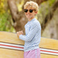 Smiling boy in The Beaufort Bonnet Company Baby Boys' and Boys' Walker's Wave Swim Shirt, sunglasses, and pink shorts holds a surfboard outdoors with trees behind him.