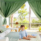 A baby wearing The Beaufort Bonnet Company's Baby Boys' Price Pool Romper in blue stripes sits on a bed with green and white linens under a canopy, with trees and a fence in the background.