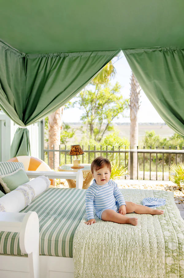 A baby wearing The Beaufort Bonnet Company's Baby Boys' Price Pool Romper in blue stripes sits on a bed with green and white linens under a canopy, with trees and a fence in the background.