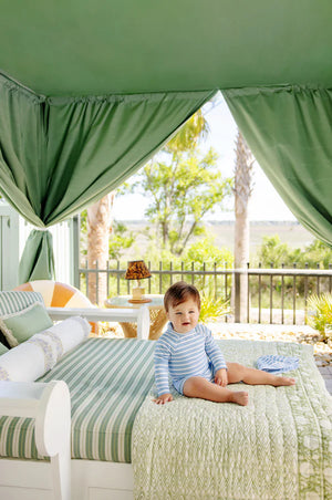 A baby wearing The Beaufort Bonnet Company's Baby Boys' Price Pool Romper in blue stripes sits on a bed with green and white linens under a canopy, with trees and a fence in the background.
