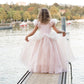 A young girl wearing the Great Pretenders Antique Princess Gown stands on a pier by the water, facing towards trees and boats in the background.