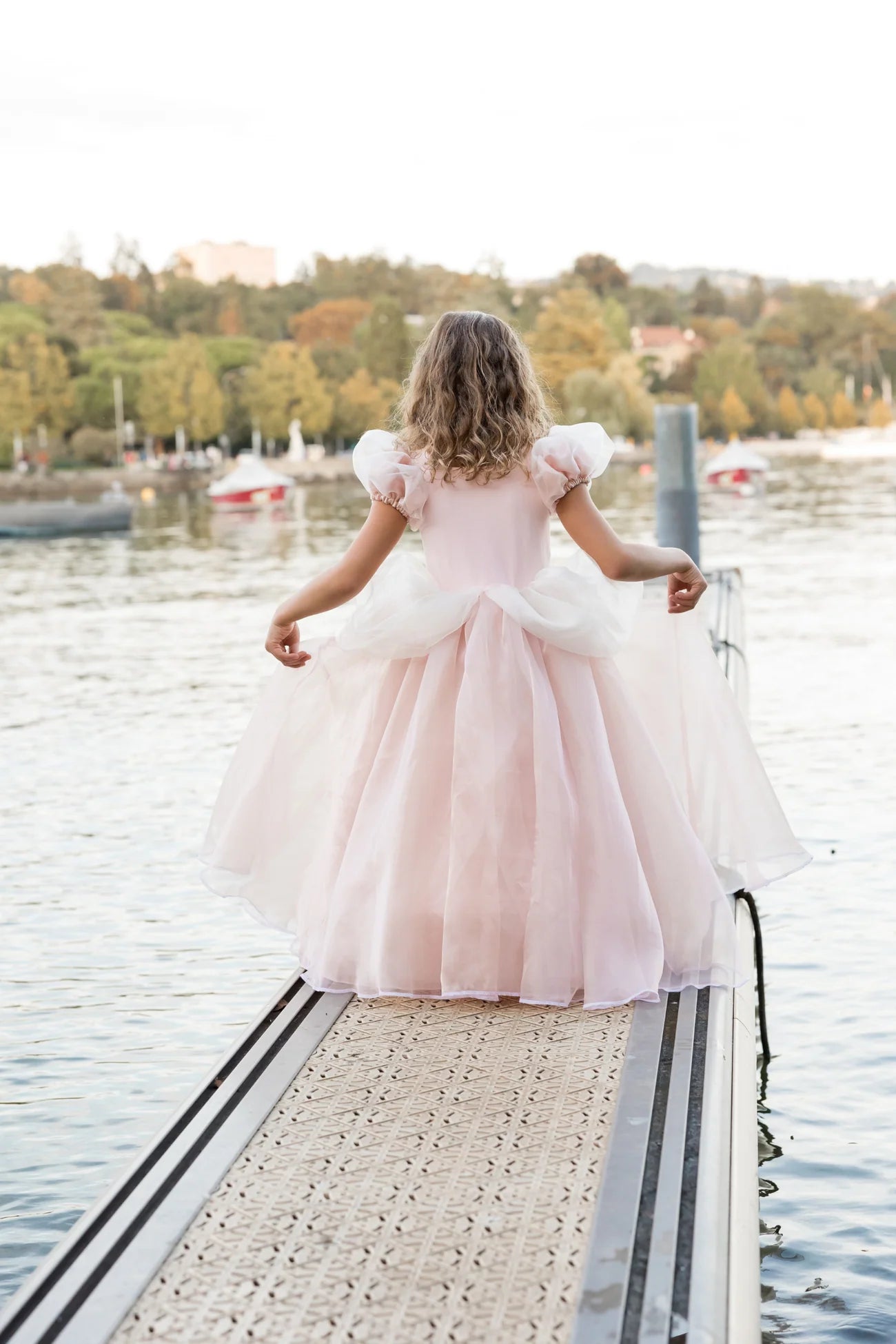 A young girl wearing the Great Pretenders Antique Princess Gown stands on a pier by the water, facing towards trees and boats in the background.