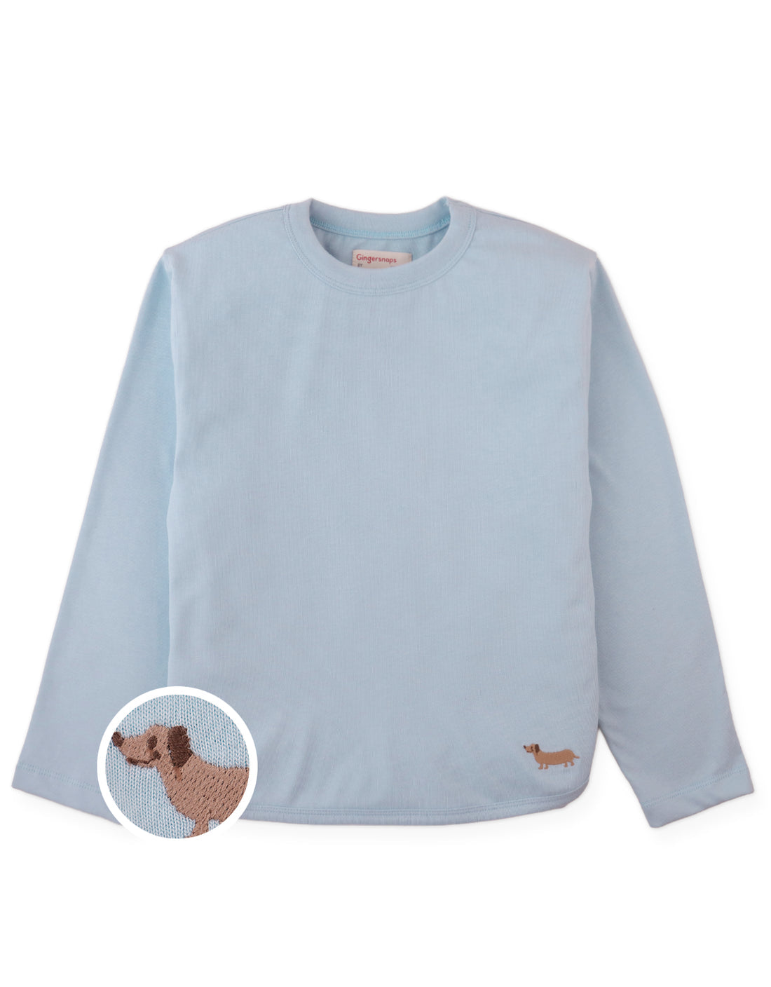 The Boys’ Teo Jersey Tee by Gingersnaps is a light blue long-sleeve shirt with subtle urban style, featuring a small embroidered brown dachshund on the lower front corner, as shown in a close-up inset.