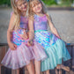 Two girls wear the Great Pretenders Bling Bling Mermaid Skirt & Hairclip Set, sitting close together on a beach, framed by sand and greenery.