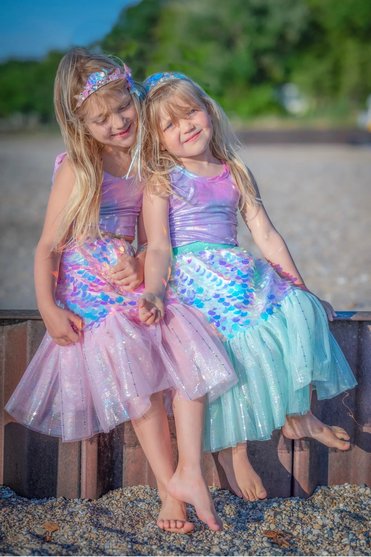 Two girls wear the Great Pretenders Bling Bling Mermaid Skirt & Hairclip Set, sitting close together on a beach, framed by sand and greenery.