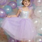 A young girl in a Great Pretenders Sequins Princess Dress with tulle skirt and a tiara smiles in front of a backdrop of colorful balloons, celebrating a special occasion.