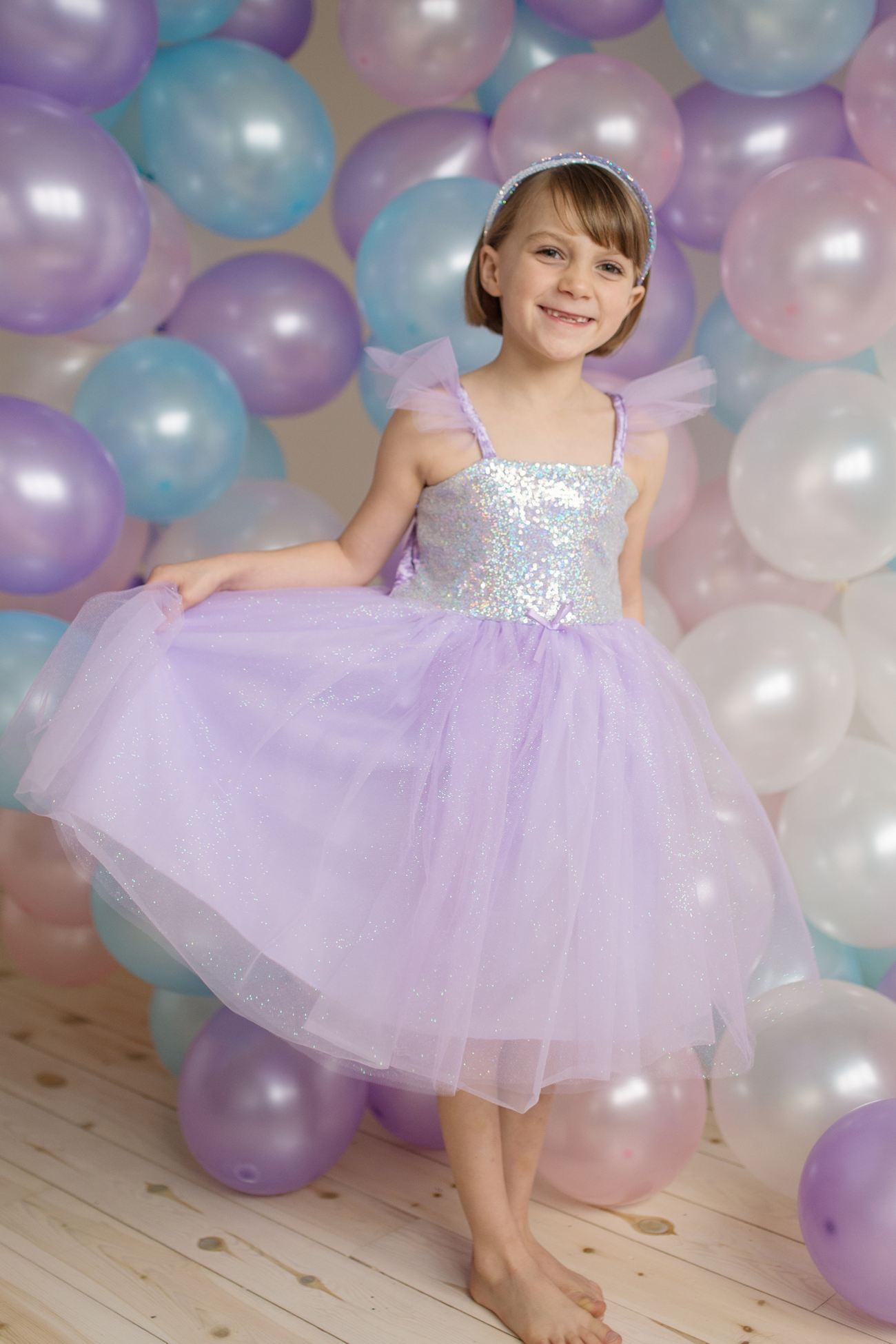 A young girl in a Great Pretenders Sequins Princess Dress with tulle skirt and a tiara smiles in front of a backdrop of colorful balloons, celebrating a special occasion.