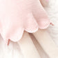 Close-up of Charlotte the Dog - Regular 20" by Cuddle + Kind, a knitted doll in a light pink scalloped dress, showing cream-colored legs on a quilted white surface with delicate wildflowers.