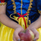 A child wearing the Great Pretenders Fairytale Princess - Snow White deluxe gown in blue, red, and yellow holds a red apple with both hands; the emphasis is on the premium quality costume and the apple rather than the child’s face.