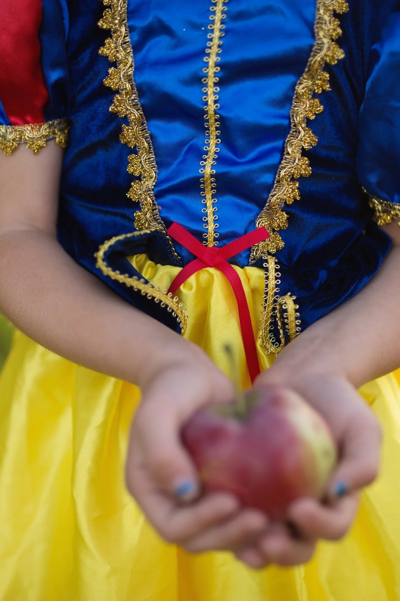A child wearing the Great Pretenders Fairytale Princess - Snow White deluxe gown in blue, red, and yellow holds a red apple with both hands; the emphasis is on the premium quality costume and the apple rather than the child’s face.