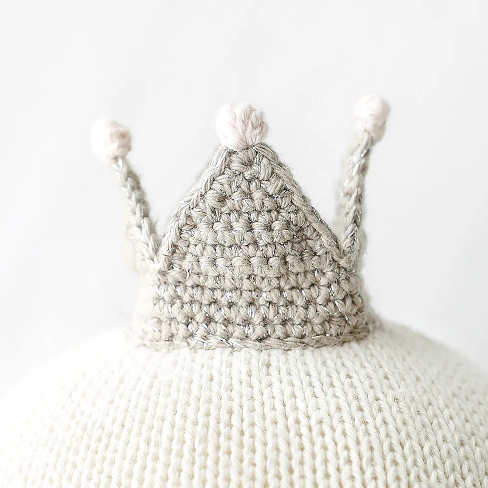 Harlow the Swan - Little 13" by Cuddle + Kind sits gracefully atop a white knitted surface, its small crocheted gray crown with three points ready to take flight against the plain background.