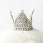 Harlow the Swan - Regular 20" by Cuddle + Kind sits gracefully on a knitted white surface, its small crocheted gray crown with three white pom-poms standing out against a plain background.