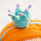 A close-up of Isla the Mermaid - Little 13" by Cuddle + Kind features a small, crocheted blue crown with purple tips on orange yarn hair, adding kindness and charm to any magical world.