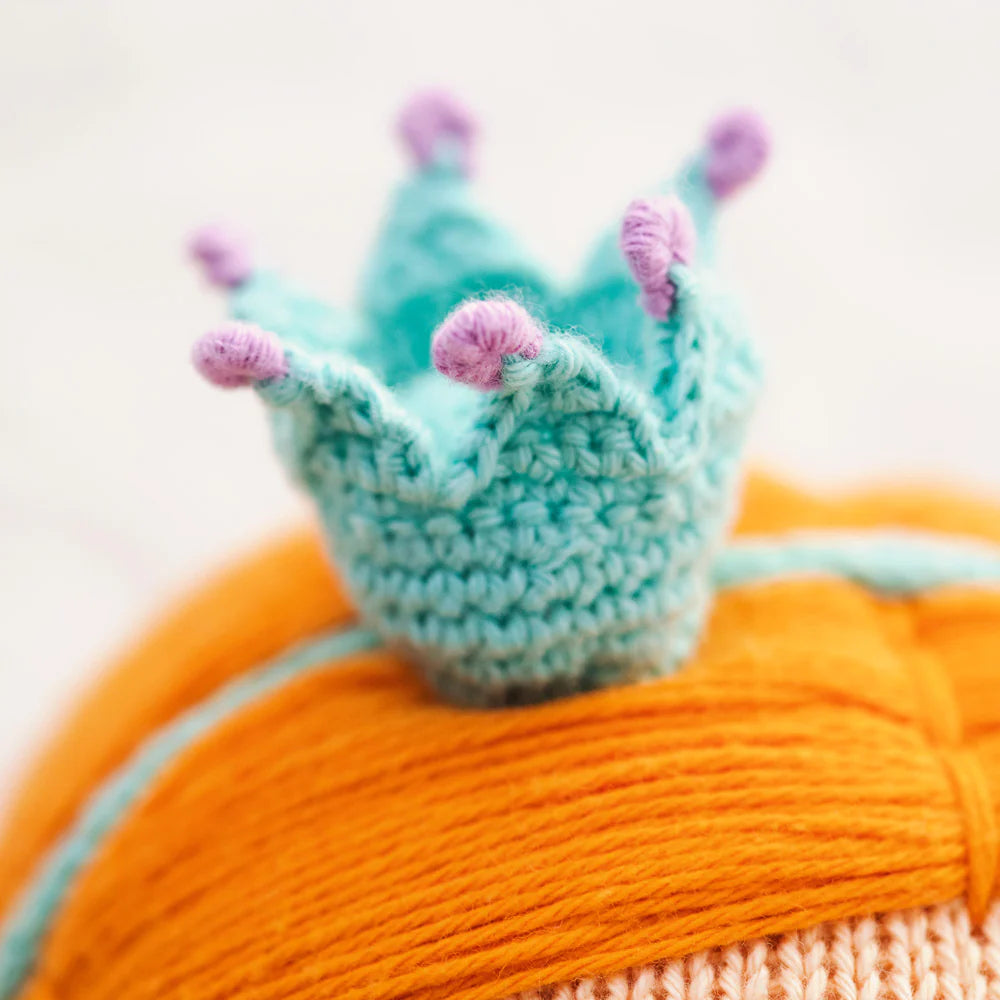 A close-up of Isla the Mermaid - Little 13" by Cuddle + Kind features a small, crocheted blue crown with purple tips on orange yarn hair, adding kindness and charm to any magical world.