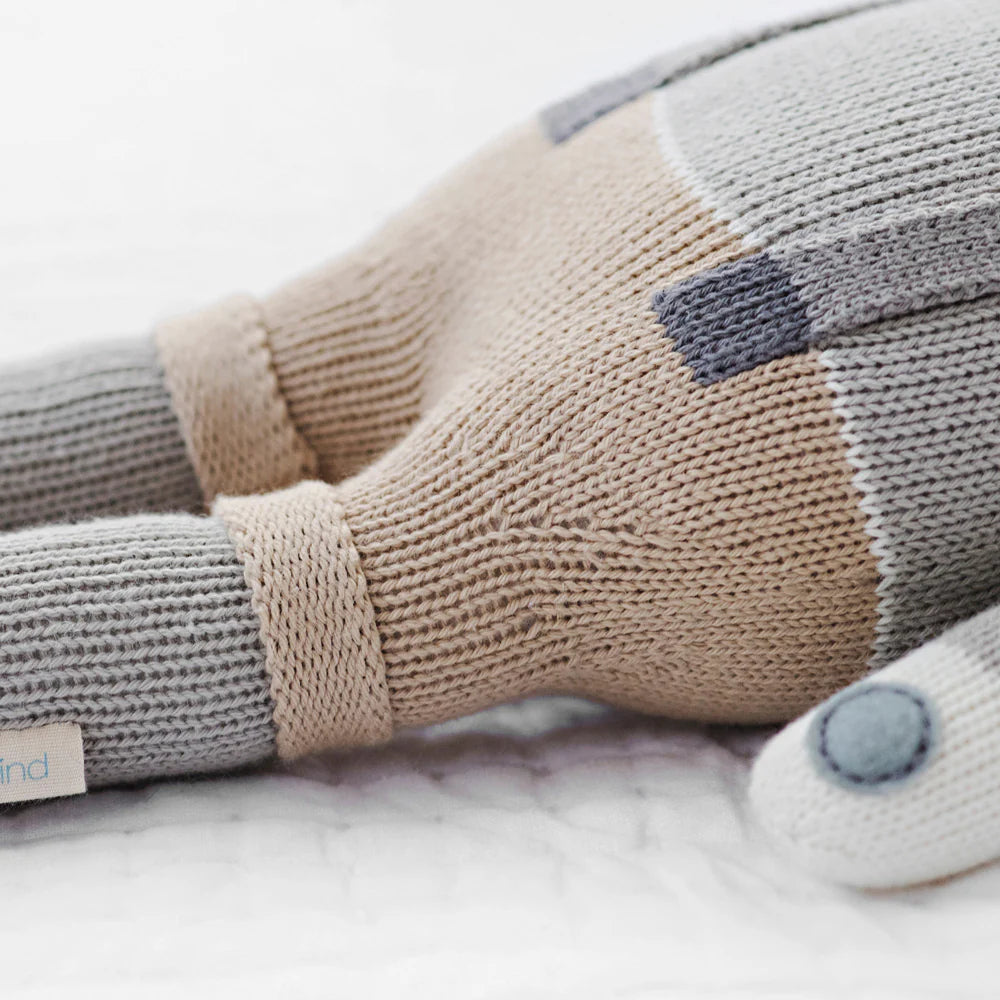 Close-up of Cuddle + Kind’s “Noah the Dog - Regular 20" plush toy, featuring beige, gray, and blue sections, resting on a white textured surface. #NoahBirthday.